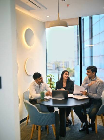 istockphoto-2193065477-612×612 Three professionals engage in a lively discussion around a round table in an office. Natural light streams in through large windows, creating a vibrant atmosphere for teamwork.