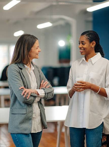 istockphoto-2204447186-612×612 Two smiling businesswomen are holding a pleasant conversation during their coffee break in a modern office environment, fostering teamwork and positive communication